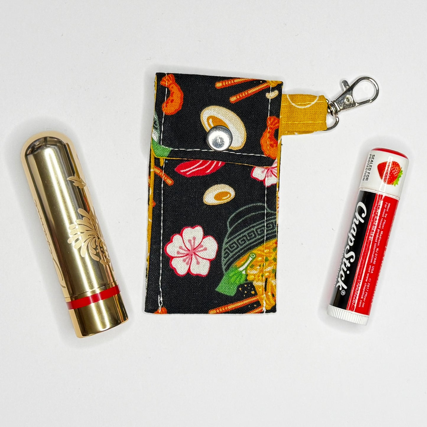 Keep your favorite lip color or balm secure and stylishly close at hand with this handmade fabric keychain pouch. Perfectly capturing a playful, retro novelty aesthetic and alternative charm, this compact accessory ensures you never lose your essentials at the bottom of your bag again.

This tailored fabric holder features a delicious, detailed ramen bowl pattern set against a stark black background, beautifully contrasted with a vibrant mustard yellow fabric loop. The print showcases savory bowls of noodle