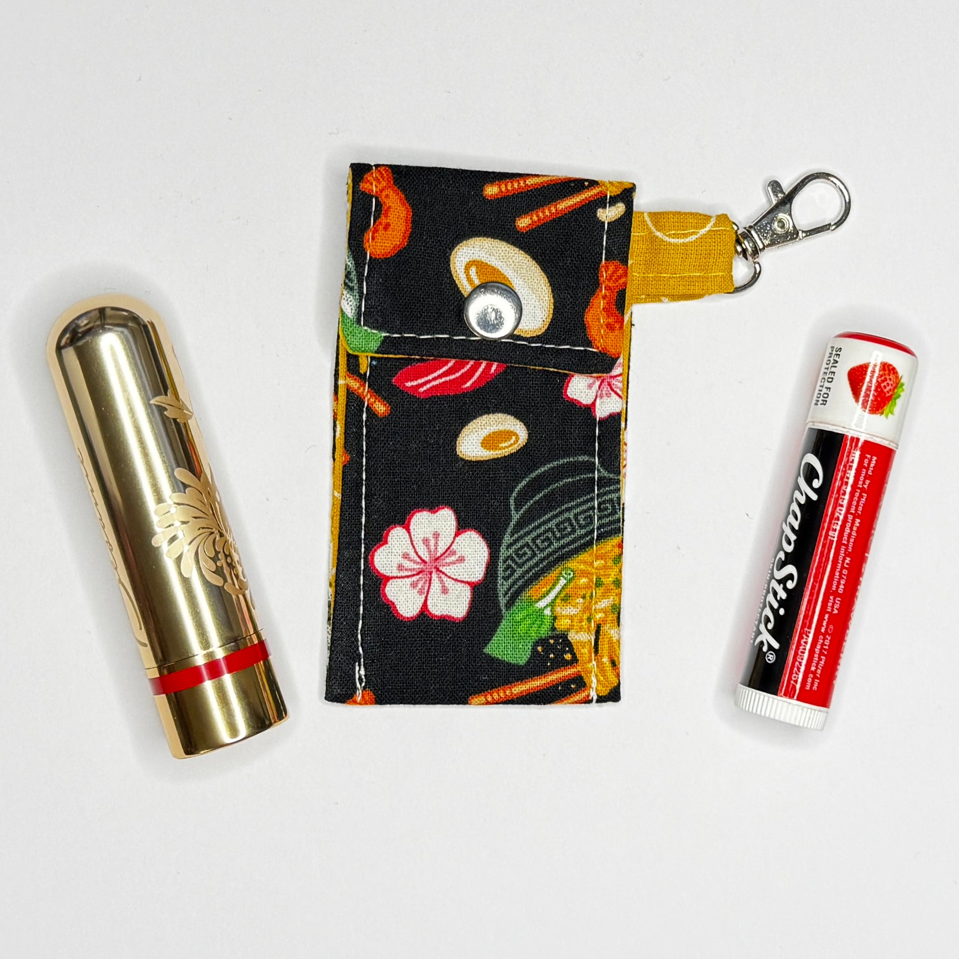 Keep your favorite lip color or balm secure and stylishly close at hand with this handmade fabric keychain pouch. Perfectly capturing a playful, retro novelty aesthetic and alternative charm, this compact accessory ensures you never lose your essentials at the bottom of your bag again.

This tailored fabric holder features a delicious, detailed ramen bowl pattern set against a stark black background, beautifully contrasted with a vibrant mustard yellow fabric loop. The print showcases savory bowls of noodle