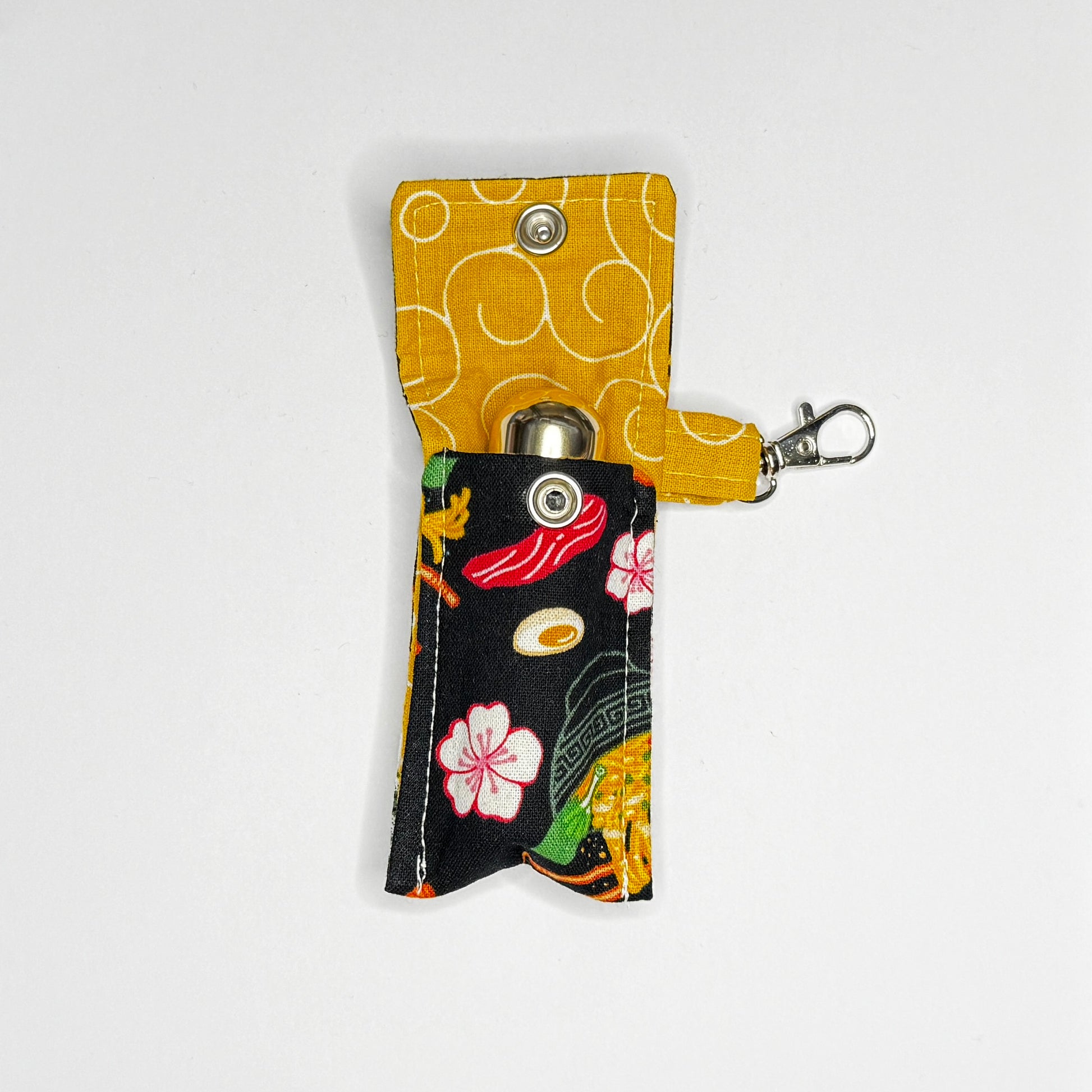 Keep your favorite lip color or balm secure and stylishly close at hand with this handmade fabric keychain pouch. Perfectly capturing a playful, retro novelty aesthetic and alternative charm, this compact accessory ensures you never lose your essentials at the bottom of your bag again.

This tailored fabric holder features a delicious, detailed ramen bowl pattern set against a stark black background, beautifully contrasted with a vibrant mustard yellow fabric loop. The print showcases savory bowls of noodle