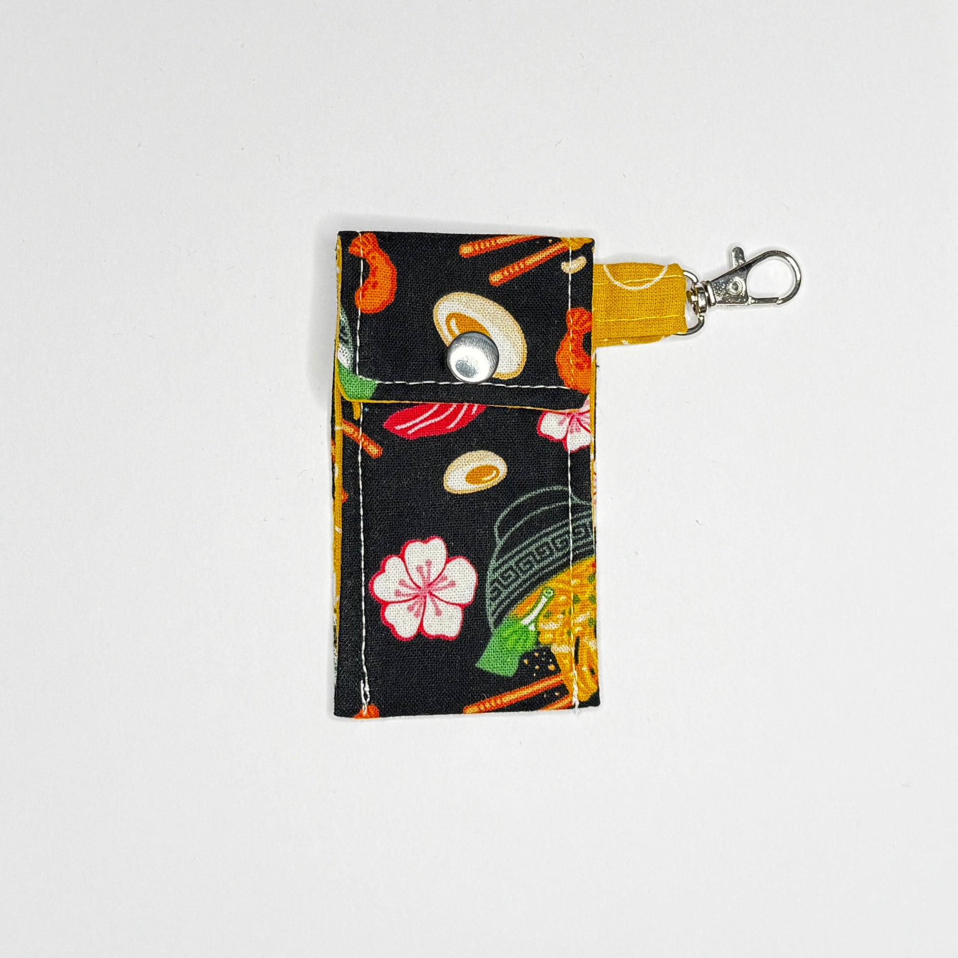 Keep your favorite lip color or balm secure and stylishly close at hand with this handmade fabric keychain pouch. Perfectly capturing a playful, retro novelty aesthetic and alternative charm, this compact accessory ensures you never lose your essentials at the bottom of your bag again.

This tailored fabric holder features a delicious, detailed ramen bowl pattern set against a stark black background, beautifully contrasted with a vibrant mustard yellow fabric loop. The print showcases savory bowls of noodle