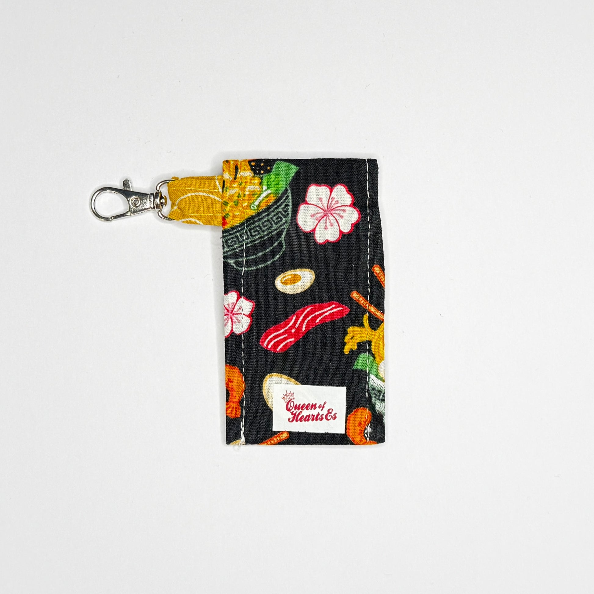 Keep your favorite lip color or balm secure and stylishly close at hand with this handmade fabric keychain pouch. Perfectly capturing a playful, retro novelty aesthetic and alternative charm, this compact accessory ensures you never lose your essentials at the bottom of your bag again.

This tailored fabric holder features a delicious, detailed ramen bowl pattern set against a stark black background, beautifully contrasted with a vibrant mustard yellow fabric loop. The print showcases savory bowls of noodle